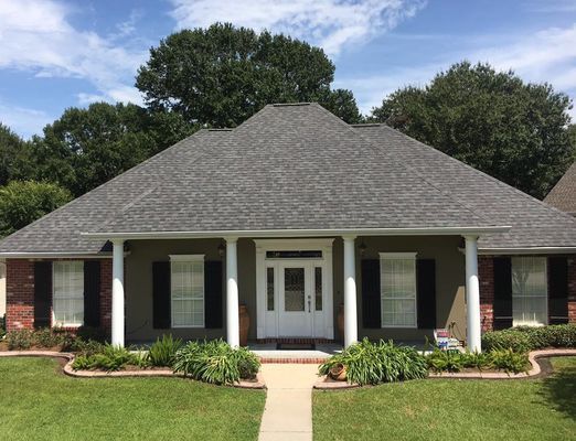 A single-story house with a grey roof, white columns, and a brick facade on a sunny day.