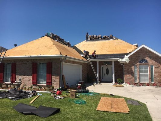 Roofers working on a residential house, replacing shingles. The roof is partially exposed plywood. The sky is clear and blue.