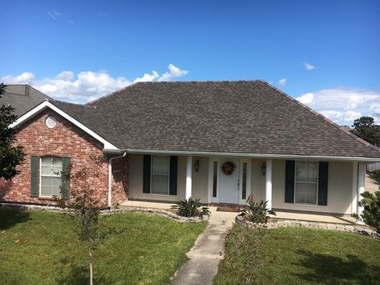 A one-story house with a brick facade and a brown shingle roof. Green lawn, white door, and a bright blue sky.