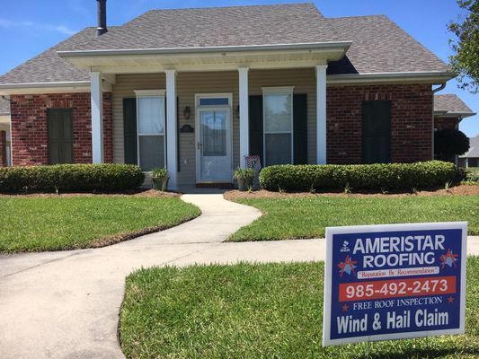 A house with a sign in the yard advertising Ameristar Roofing, specializing in wind and hail claims. The house has a beige exterior with brick accents and a green lawn.