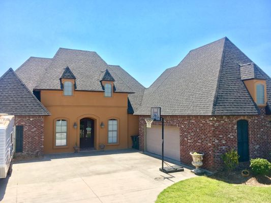 A two-story house with a brown roof, tan stucco, red brick accents, and a basketball hoop in the driveway.