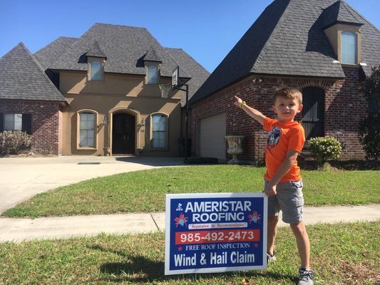 Young boy points to a house with a new roof, standing in front of an Ameristar Roofing sign on a lawn.