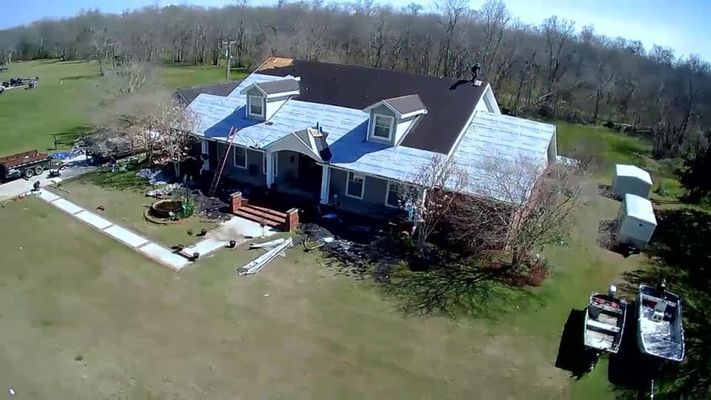 Aerial view of a house with a partially replaced roof, surrounded by a grassy yard and trees. Boats sit in the foreground.