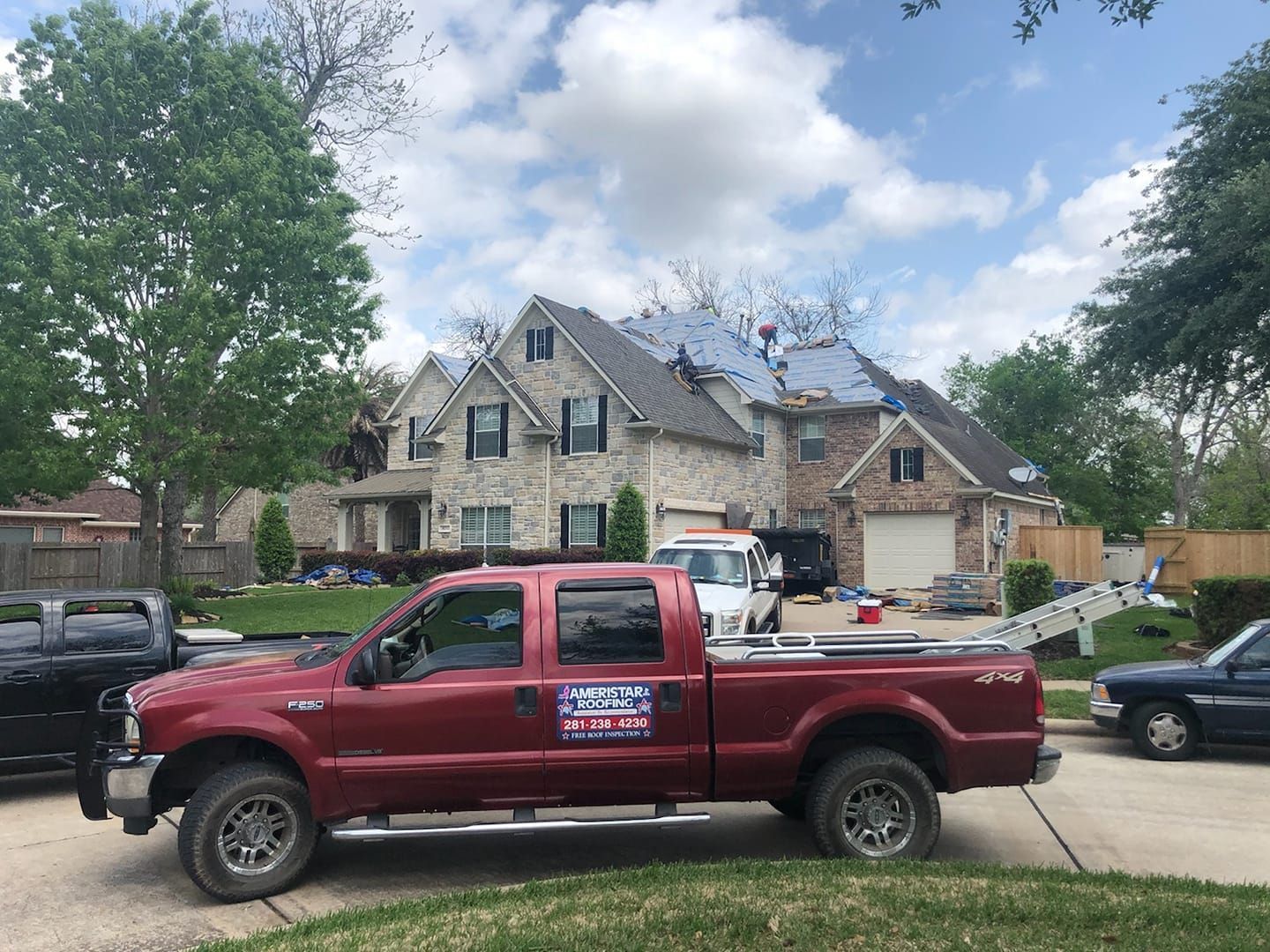 Red pickup truck parked in front of a two-story house undergoing a roof replacement. A white truck and other vehicles are in the driveway.
