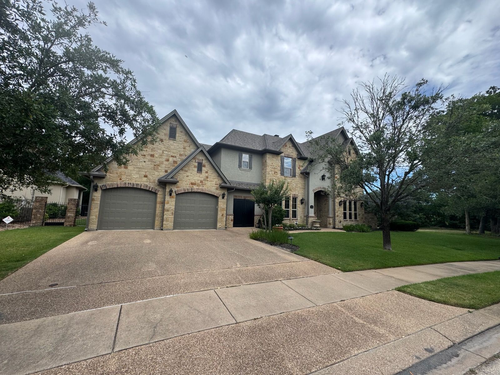 Two-story stone house with a three-car garage, green lawn, and overcast sky.