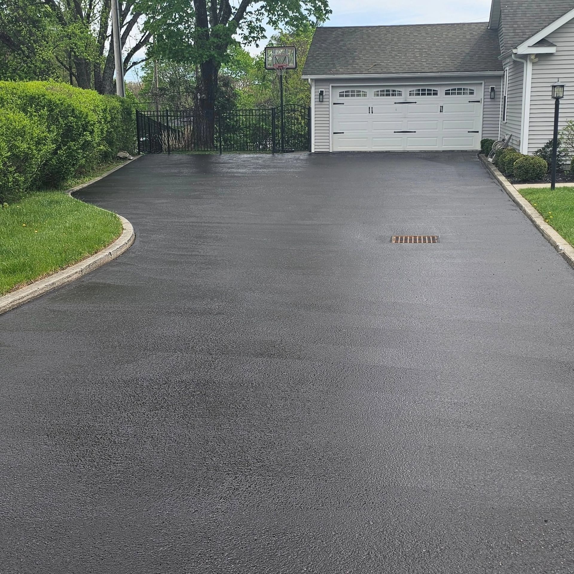 Freshly paved black driveway leading to a white garage, bordered by green shrubs and trees.