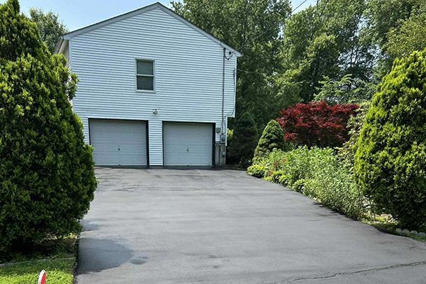 Learn More About Paving and Sealcoating A two-story house with white siding and a two-car garage at the end of a paved driveway, flanked by green bushes.