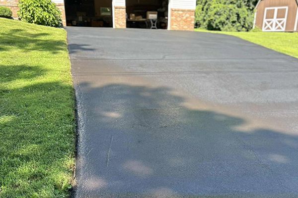 Learn More About Paving and Sealcoating A freshly sealed, dark gray asphalt driveway leads toward a residential garage next to a green lawn.