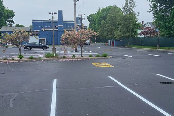 Learn More About Paving and Sealcoating An empty parking lot with marked spaces in front of a blue commercial building with a smokestack under a cloudy sky.
