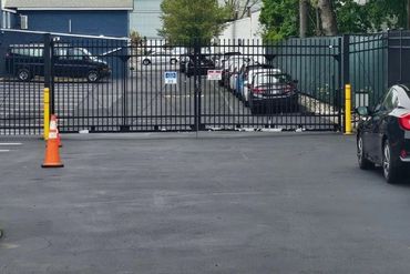 Black metal gate blocking a parking lot driveway, with orange traffic cones and parked cars behind it