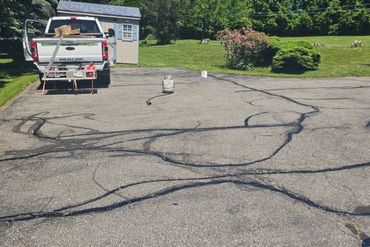 Cracked asphalt driveway with black sealant lines, a white pickup truck, and a small house in the background