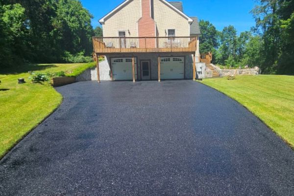 Fresh black asphalt driveway leading to a beige house with a wooden deck and two garage doors