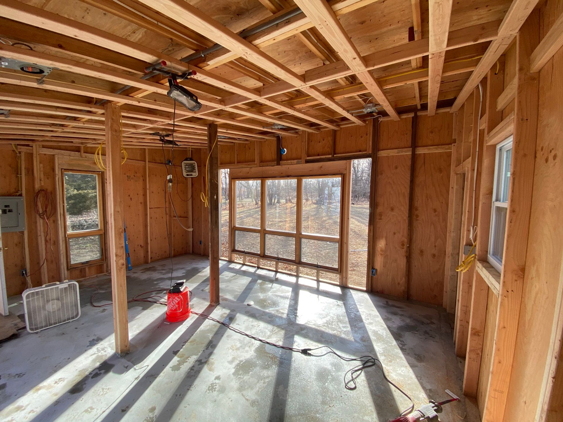 Unfinished wooden room with exposed studs, ceiling joists, and sunlight streaming through large windows.