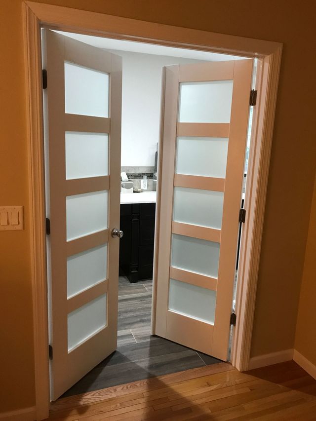 Open double glass doors leading to a kitchen, with warm wood floors and beige walls.