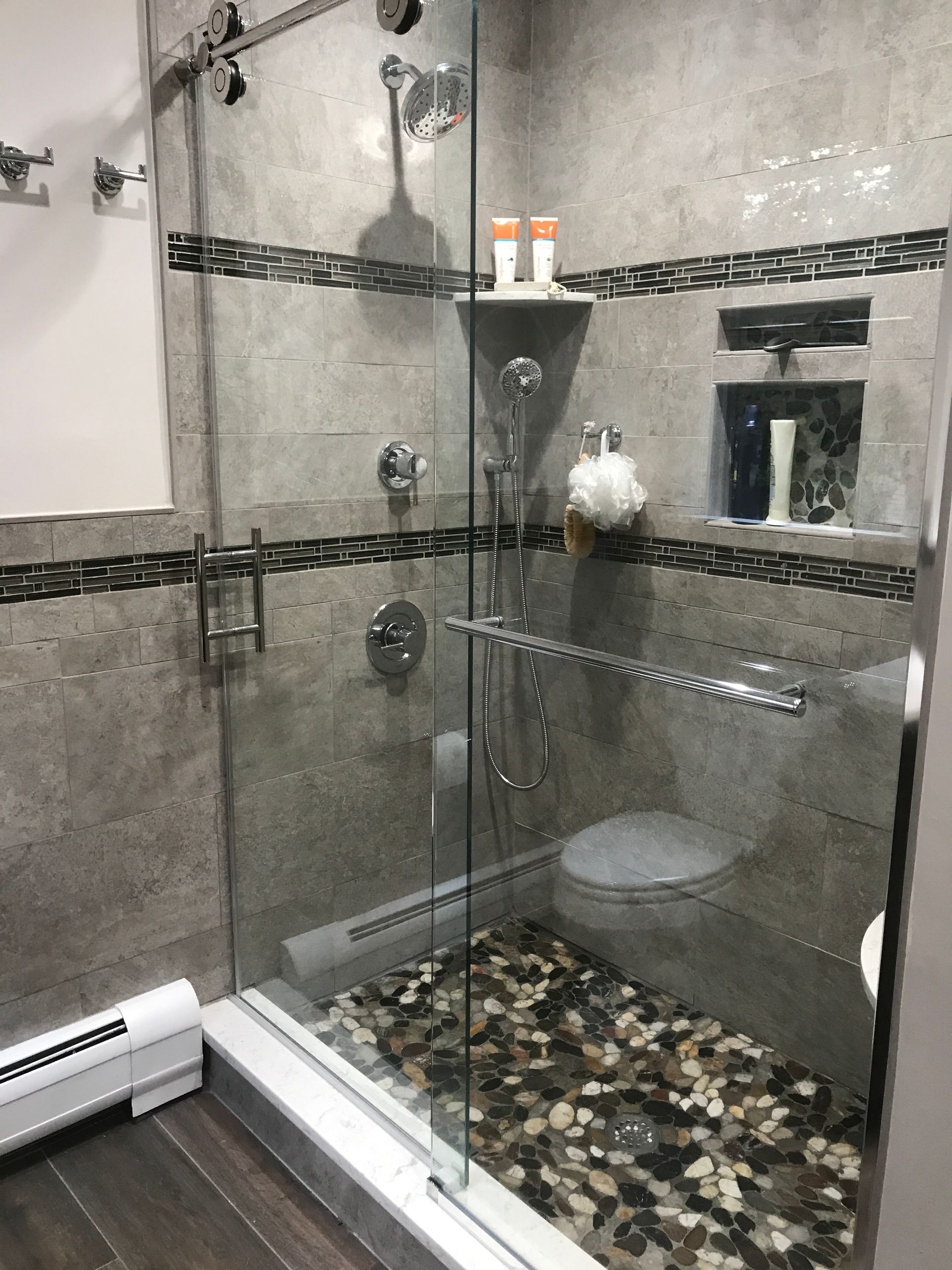 Corner glass shower with mosaic tile floor, gray walls, and a small shelf holding toiletries