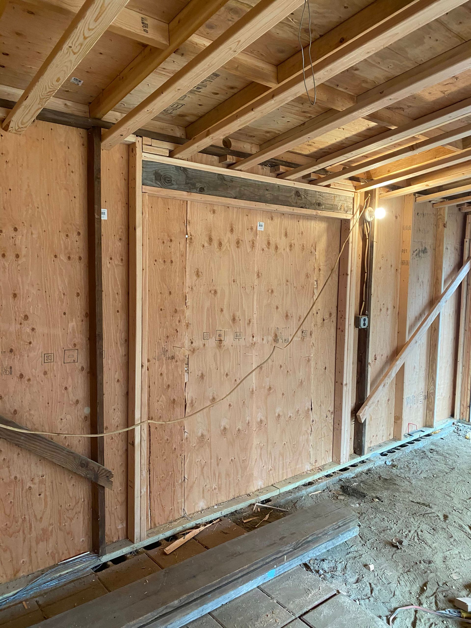 Unfinished wooden interior wall framing with plywood sheathing and a dirt floor under construction