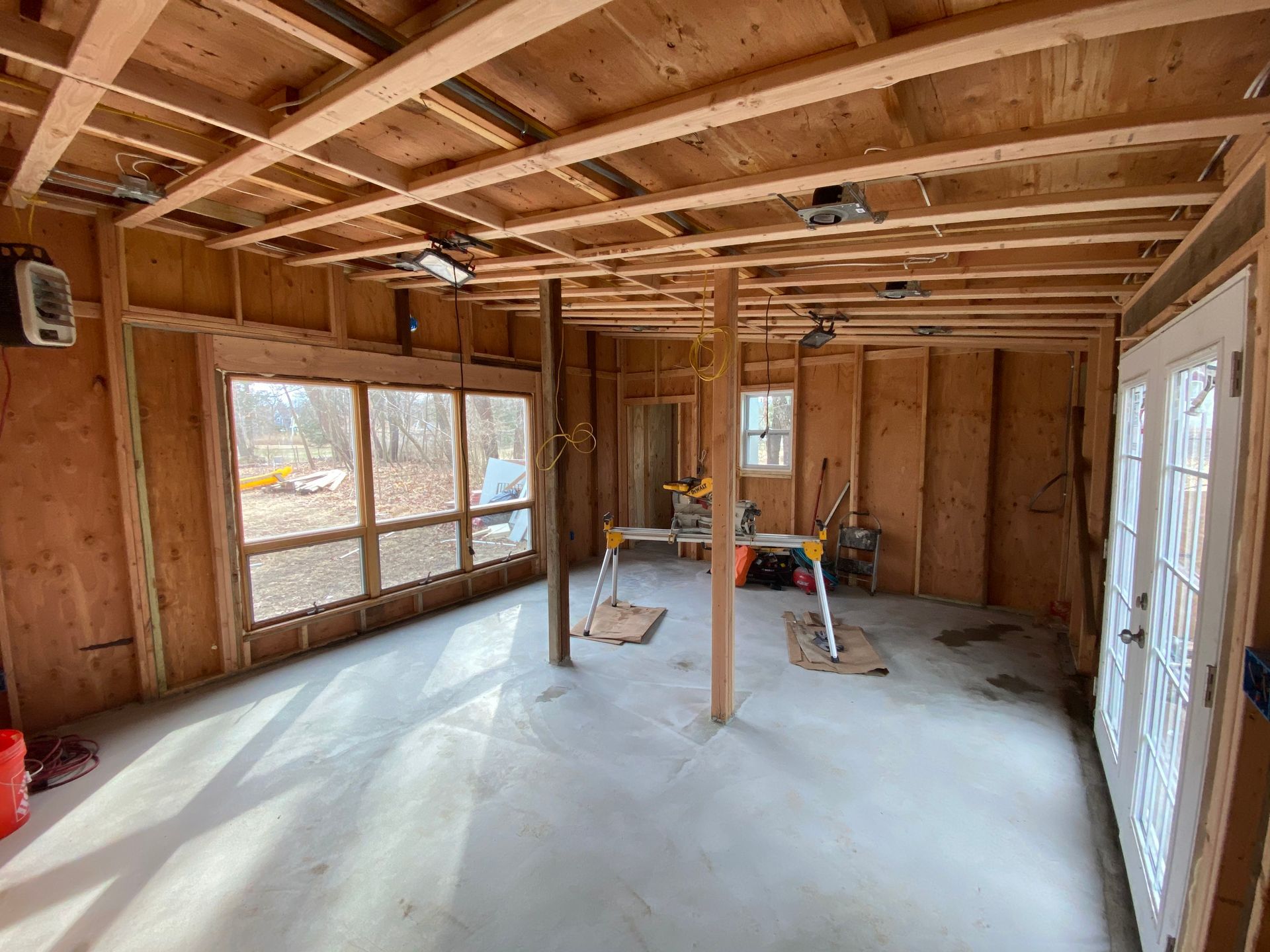Unfinished room with exposed wood framing, concrete floor, large windows, and construction tools inside.