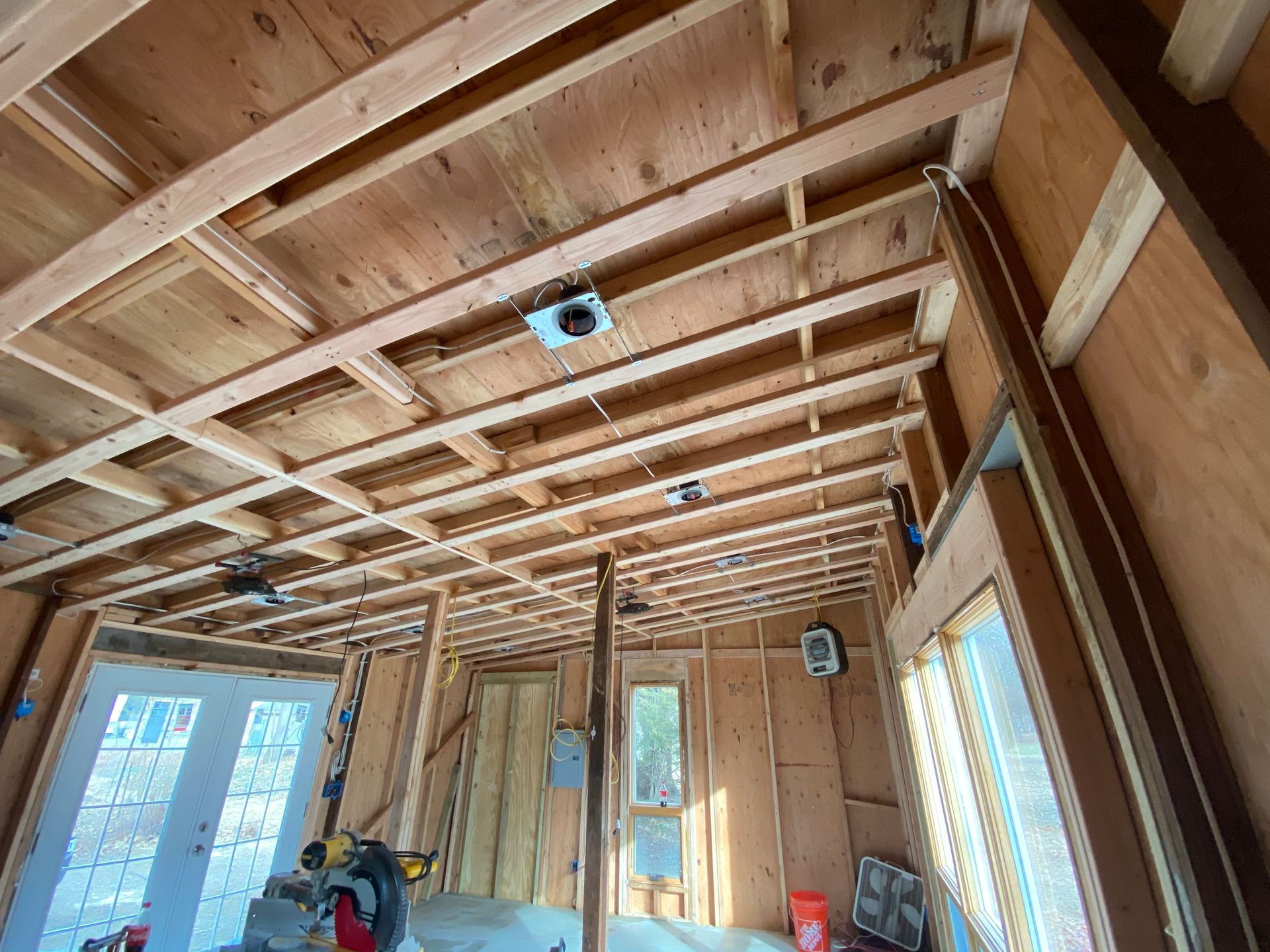 Wood-framed interior under construction with exposed ceiling beams, windows, and a ladder nearby