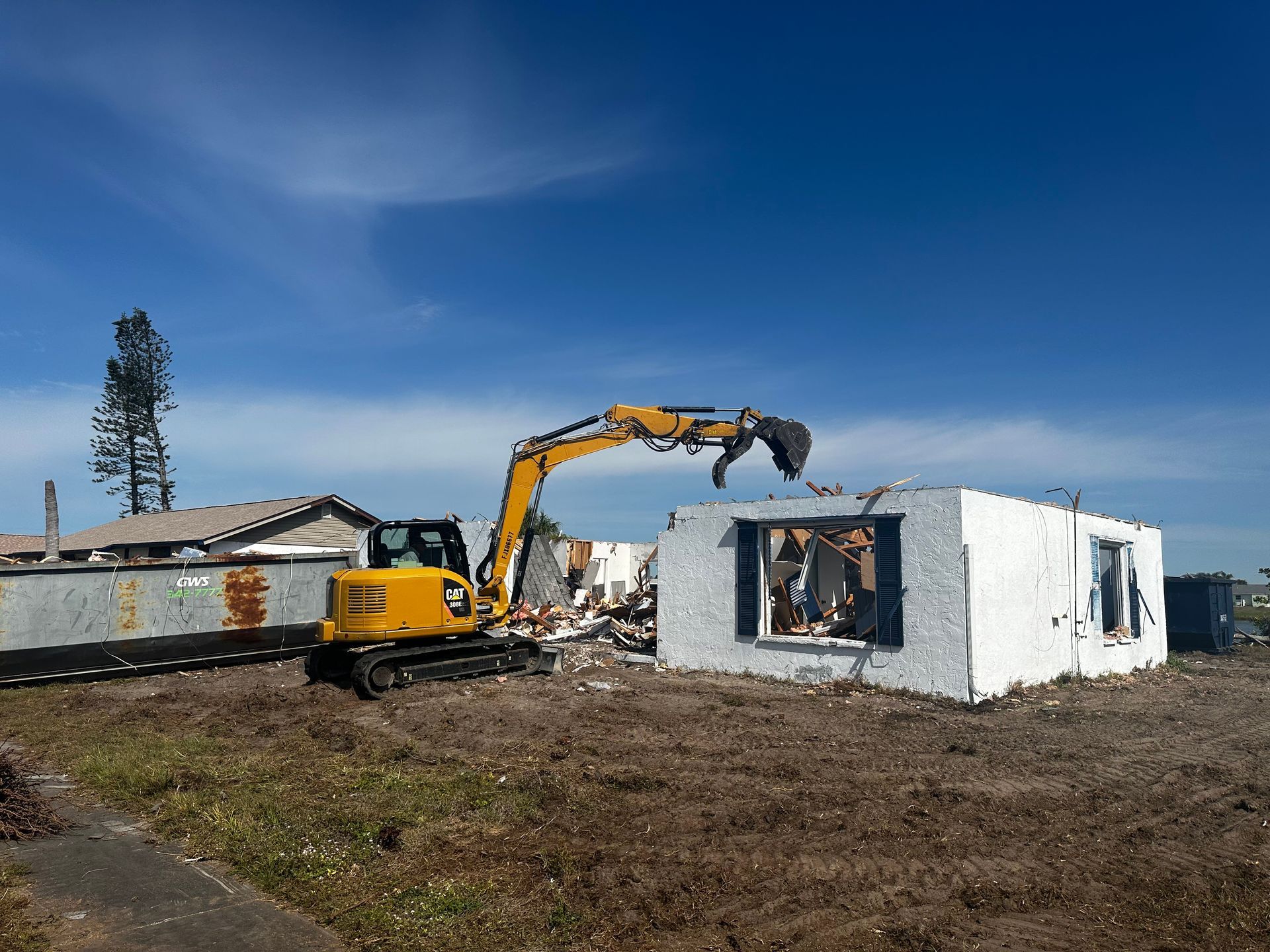 Excavator demolishing a white house under a blue sky on a muddy lot