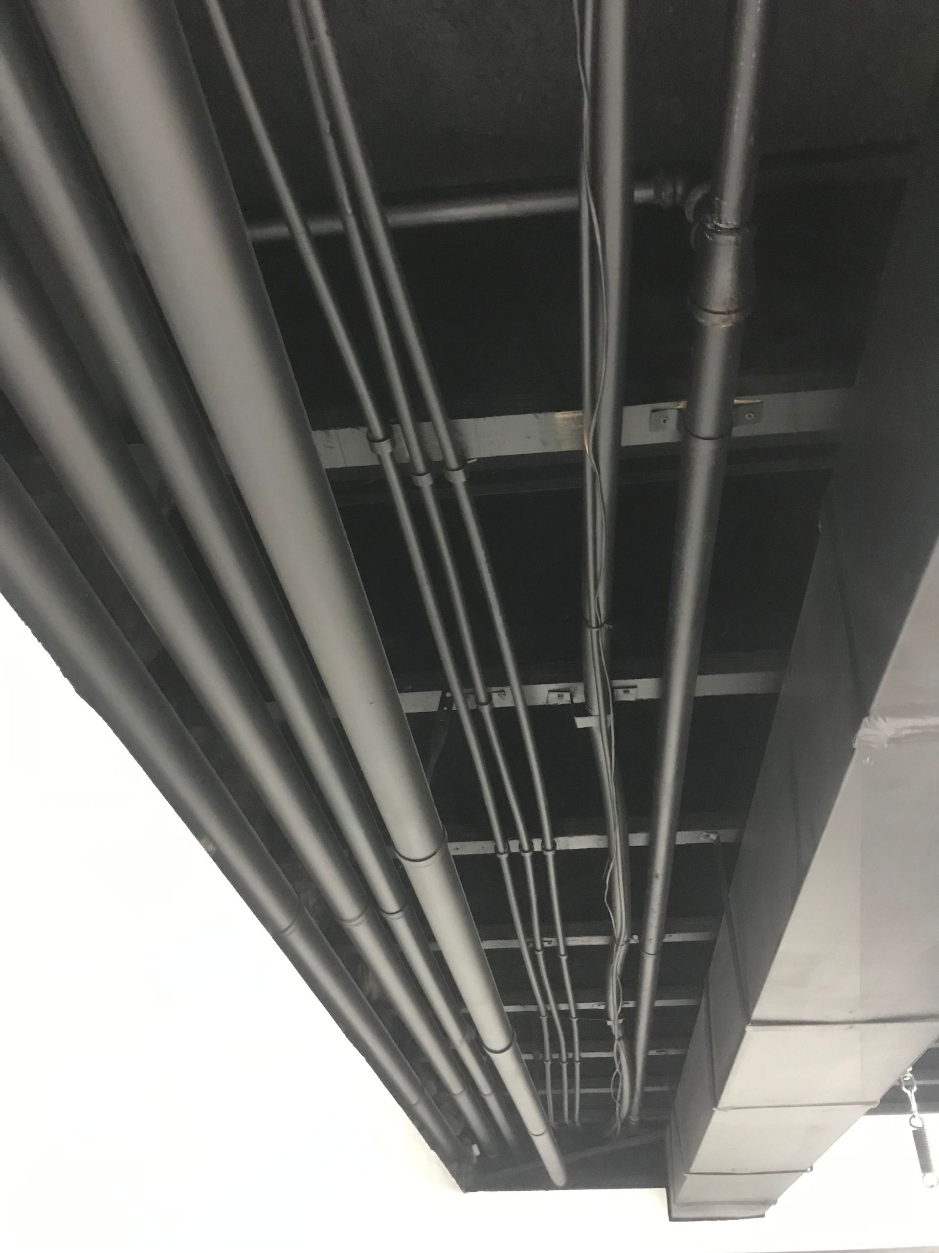 Underside of a bridge or overpass with exposed beams and utility pipes, shot upward against a bright sky