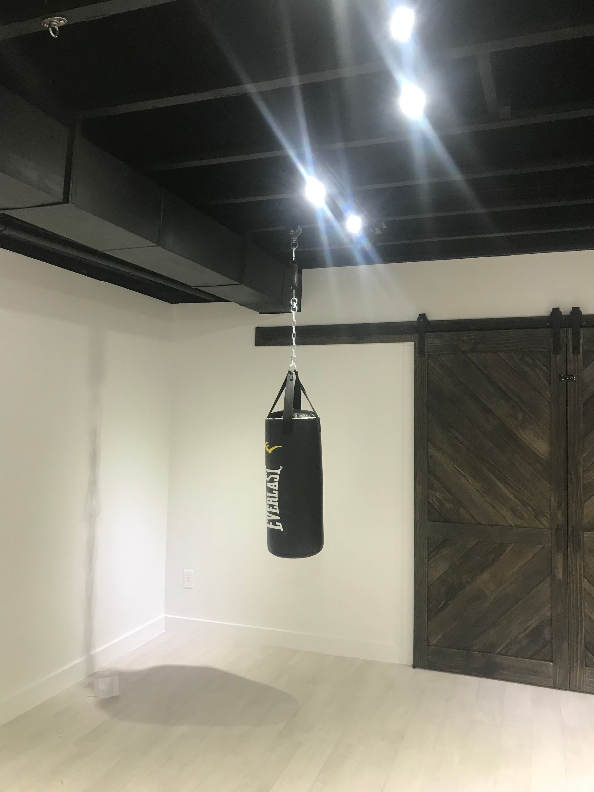 Black punching bag hanging in a minimalist gym room with white walls and wooden double doors.