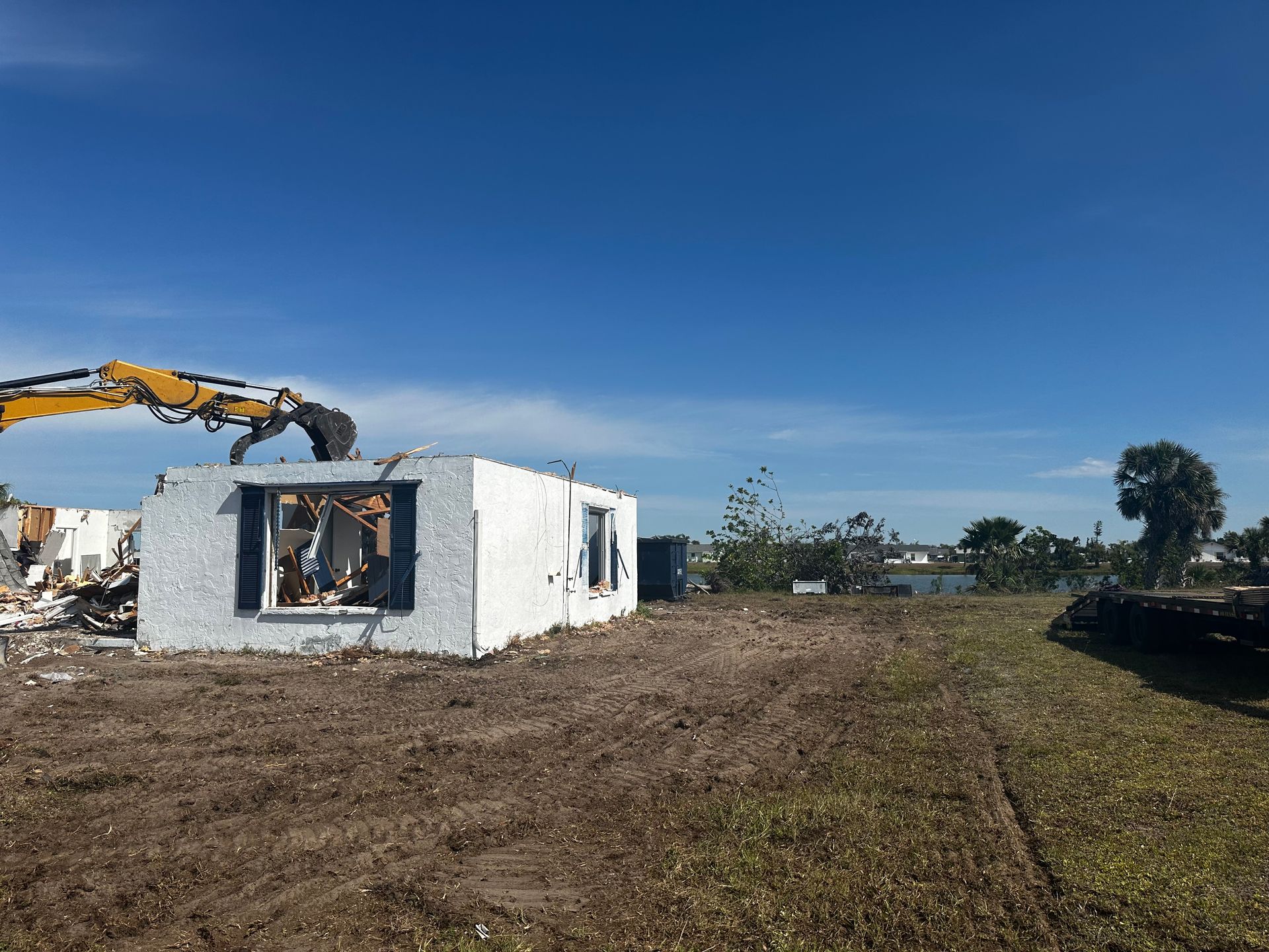 Excavator demolishing a small white house on a dirt lot under a clear blue sky