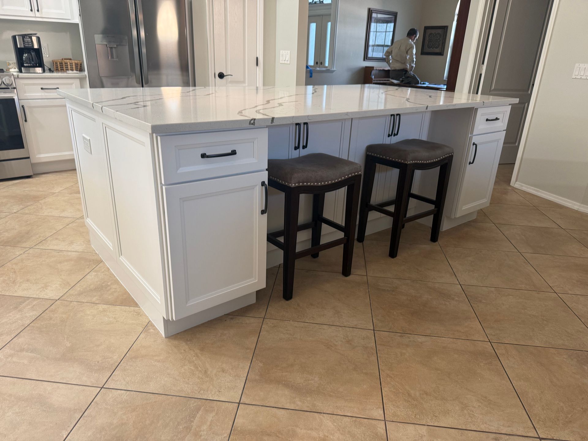 Bright kitchen with a white island, two black stools, and beige tile flooring.