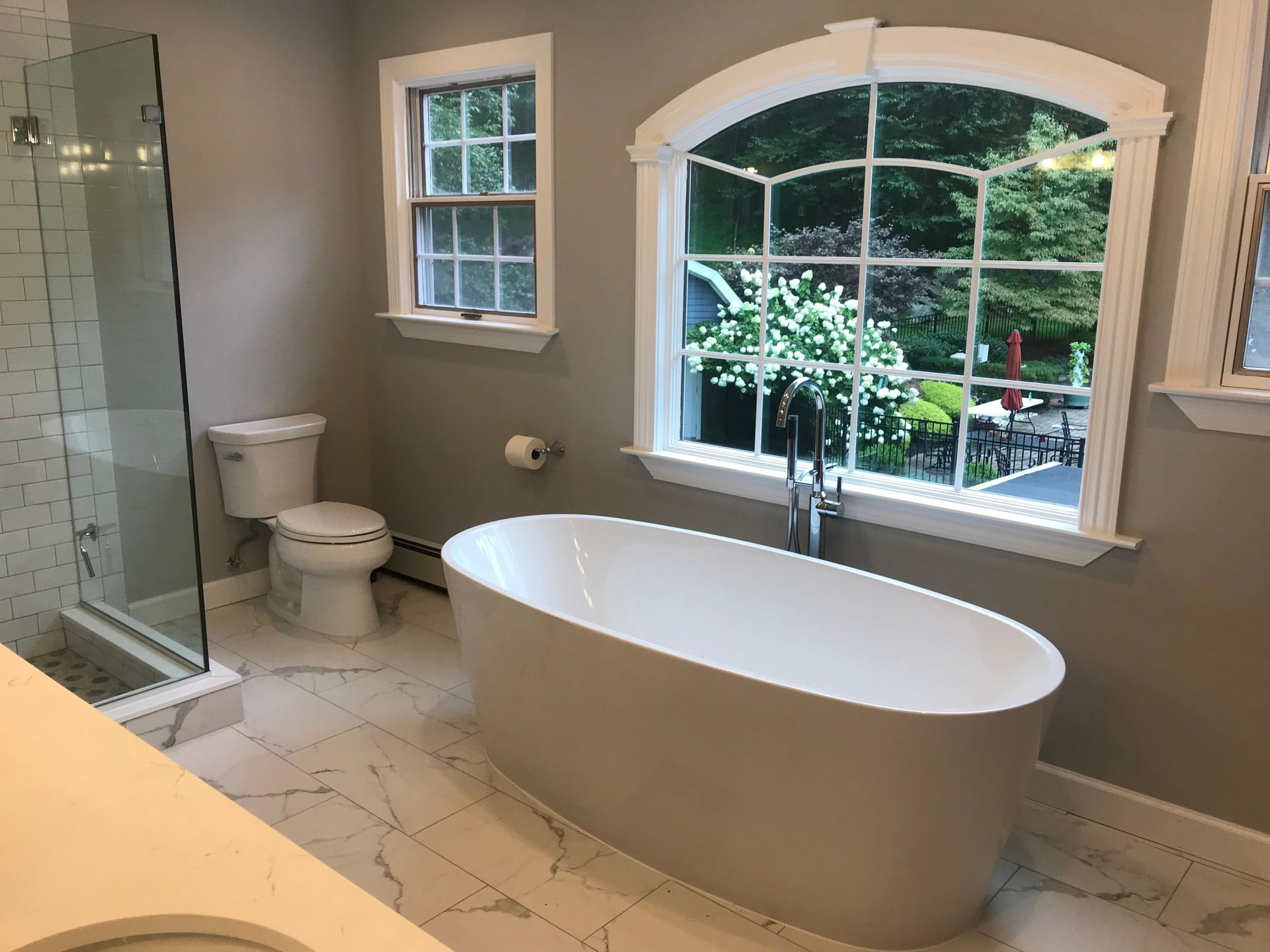 Modern bathroom with a freestanding tub, glass shower, toilet, and large arched window overlooking greenery