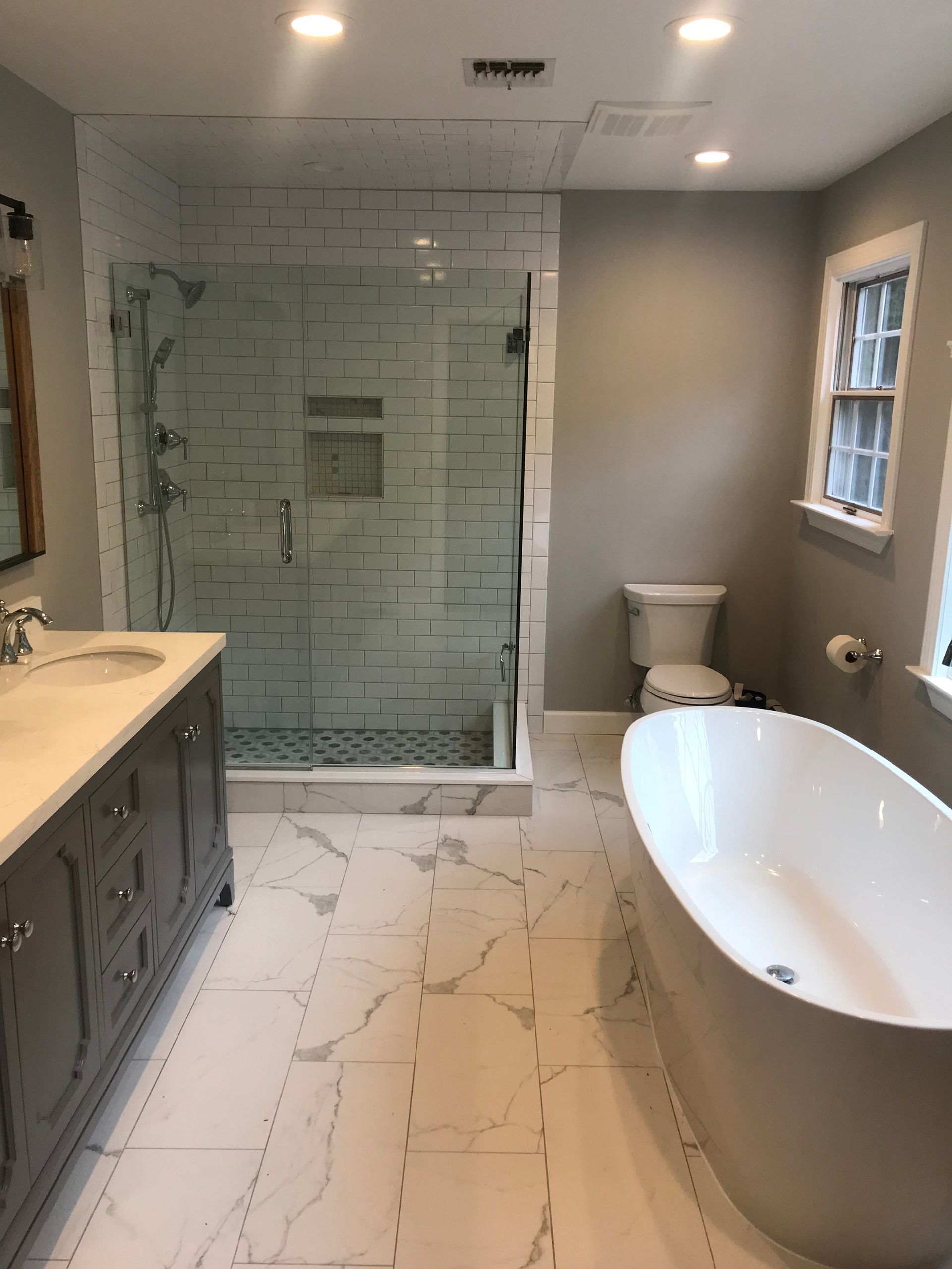 Modern bathroom with glass shower, white marble floor, vanity, toilet, and freestanding tub