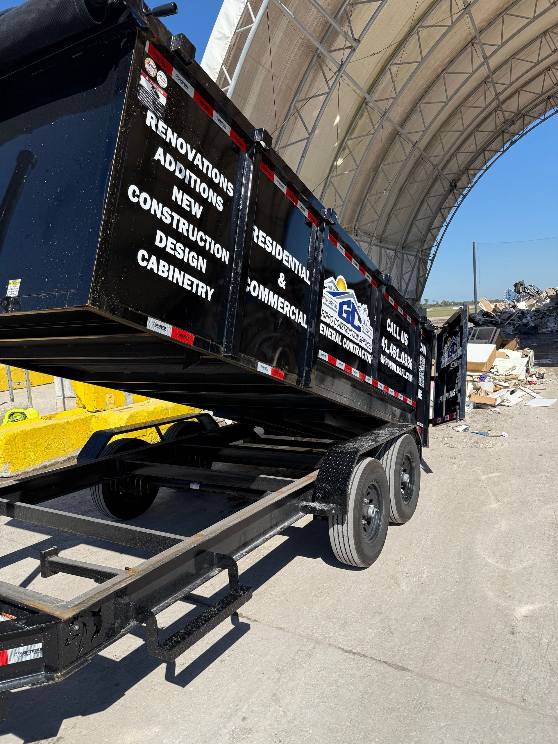 Black dump trailer with white lettering under a large arched canopy on a dusty roadside