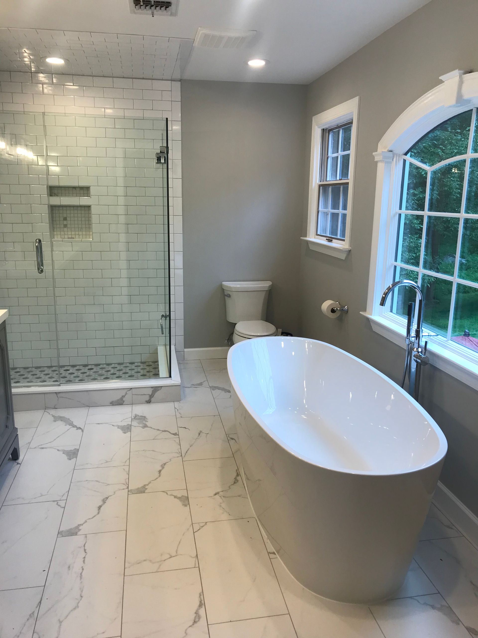 Modern bathroom with freestanding tub, glass shower, white tile, and arched window