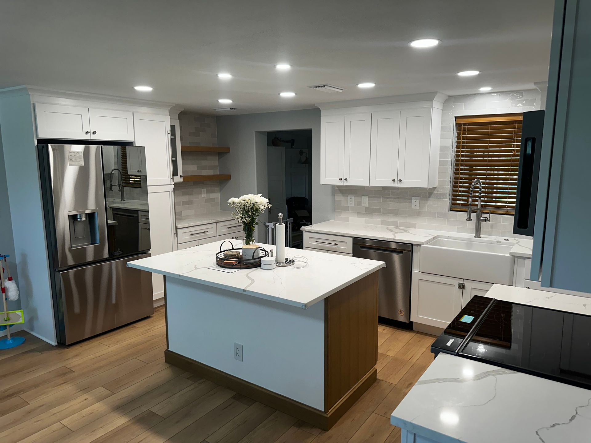 Modern white kitchen with stainless fridge, island, and wood floors, brightly lit by recessed ceiling lights