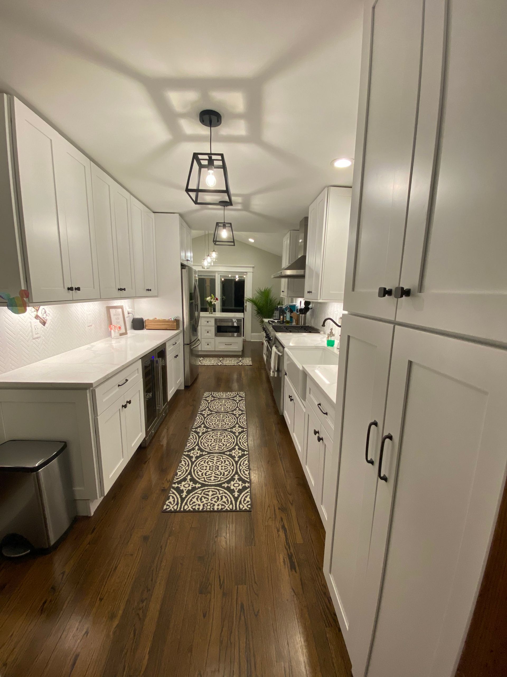 Narrow white kitchen with wood floors, patterned runner, and pendant lights leading to a doorway.