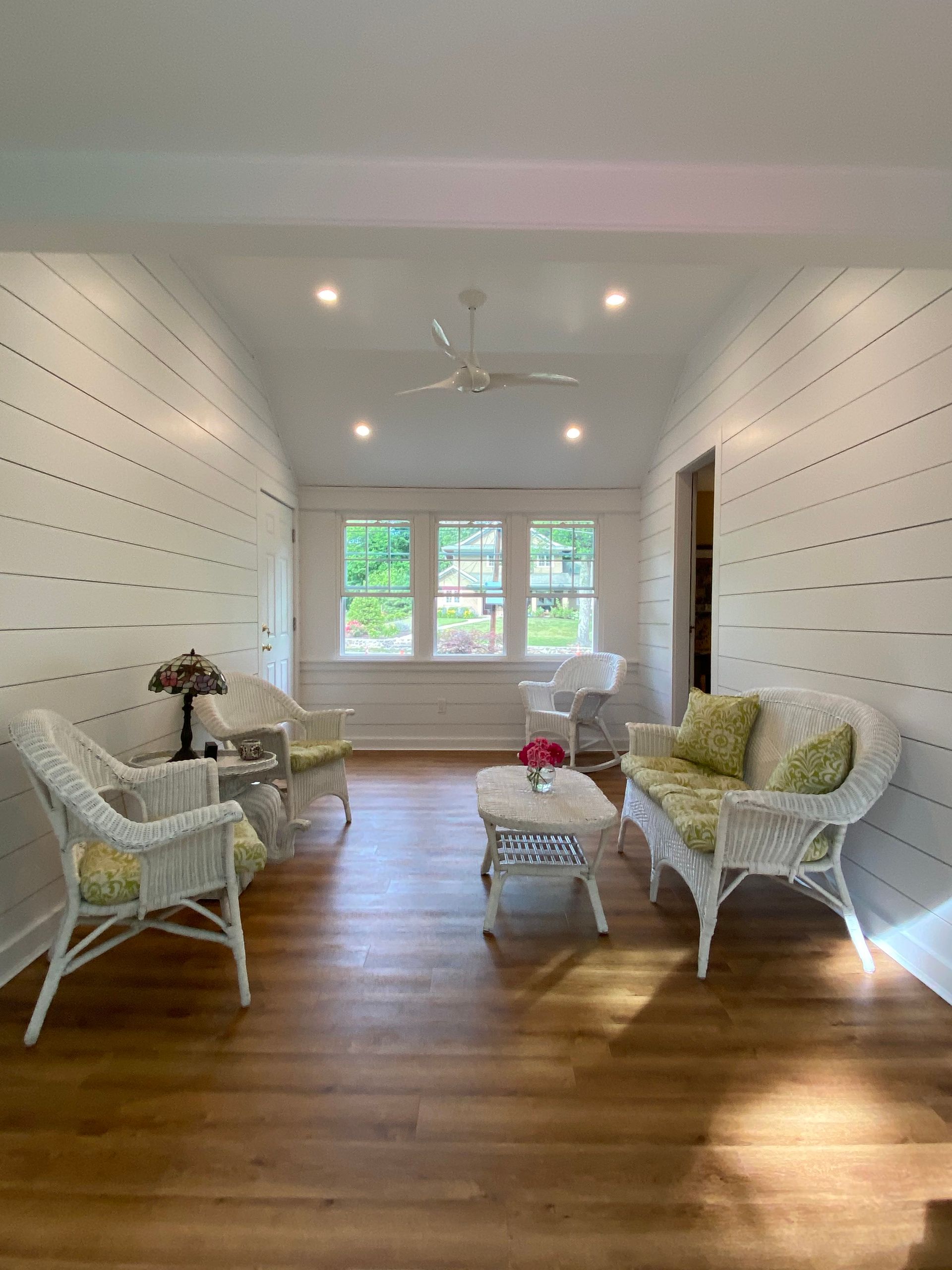 Bright sunlit living room with wicker chairs, coffee table, wood floors, and a window at the far end