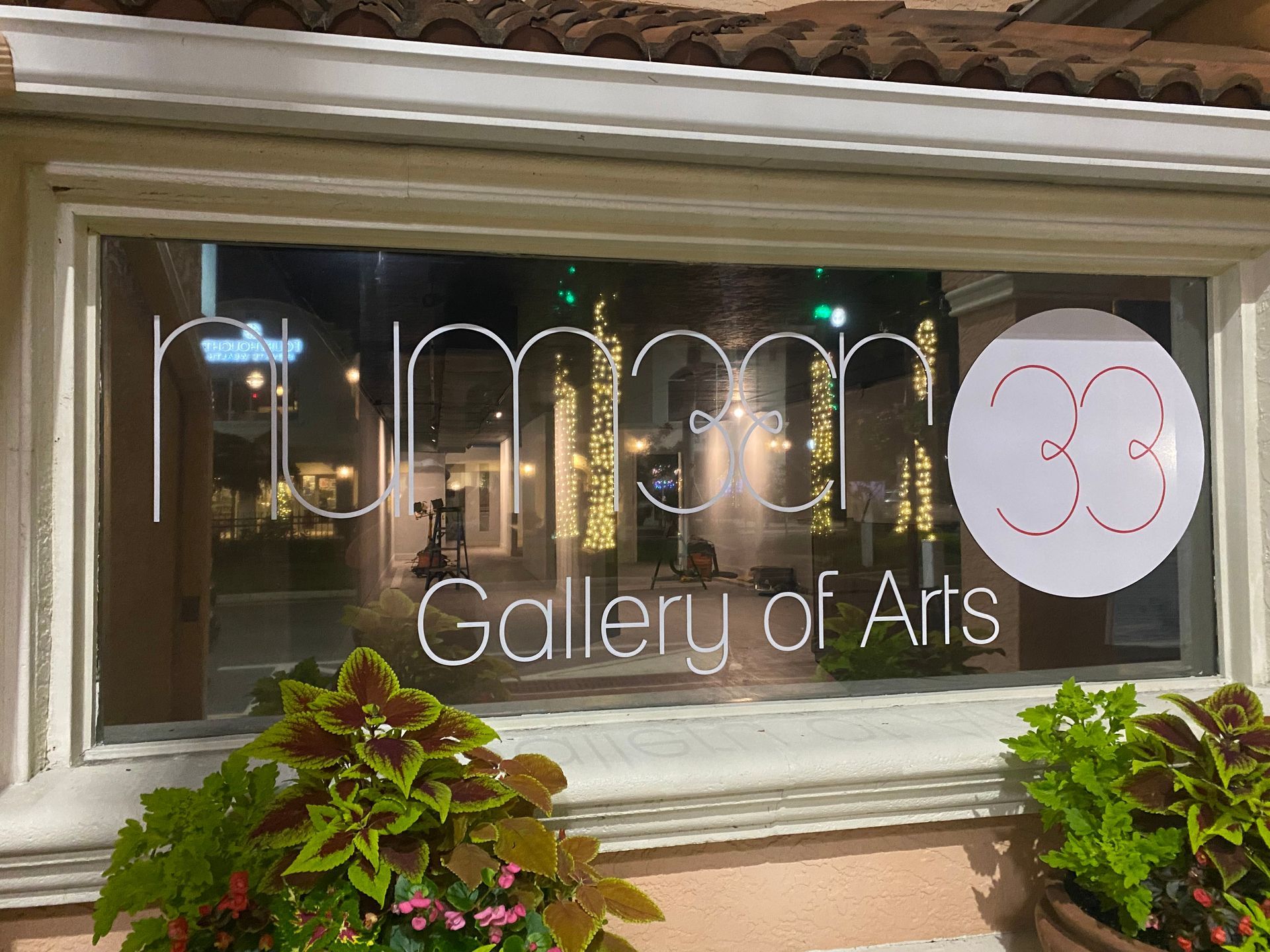 Wine bar window sign reading “lme 30 Gallery of Arts,” with plants below