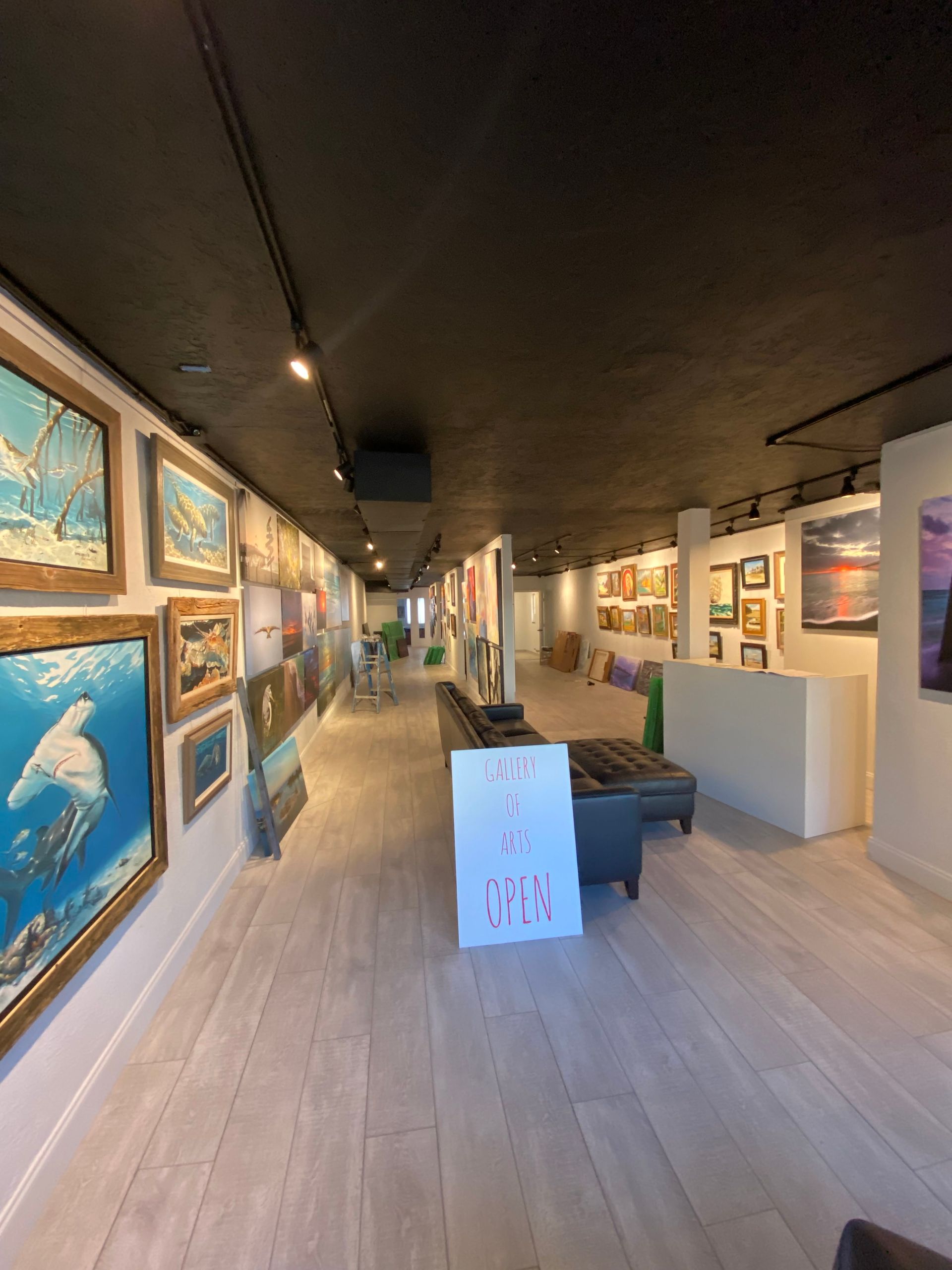 Art gallery interior with framed paintings, dark ceiling, and a central bench on a light wood floor.