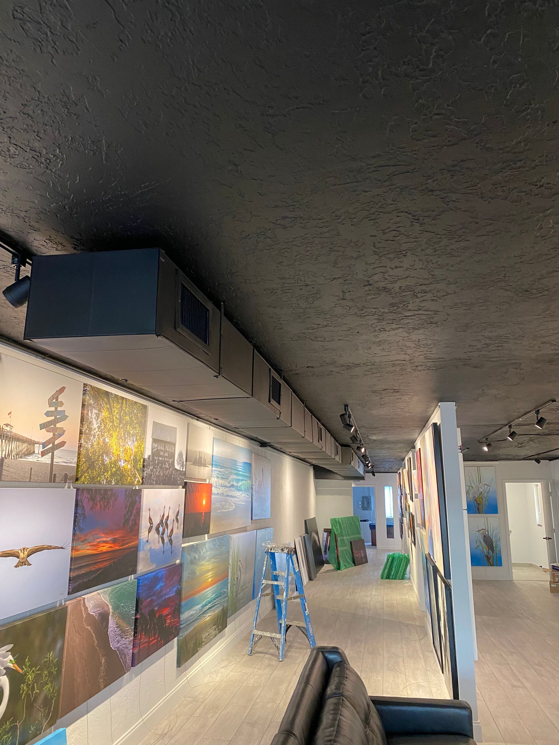 Gallery hallway with colorful wall art, concrete ceiling, and stools along a narrow corridor