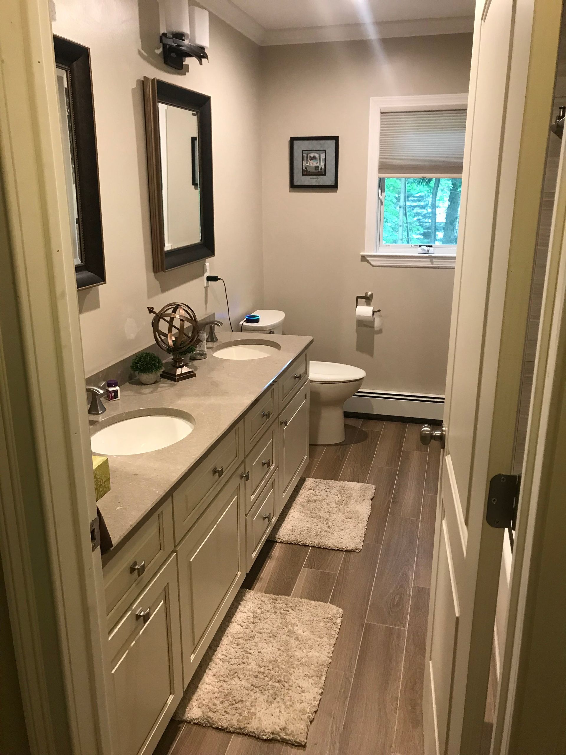 Narrow beige bathroom with double vanity, wall mirror, rug runner, and window at the end.