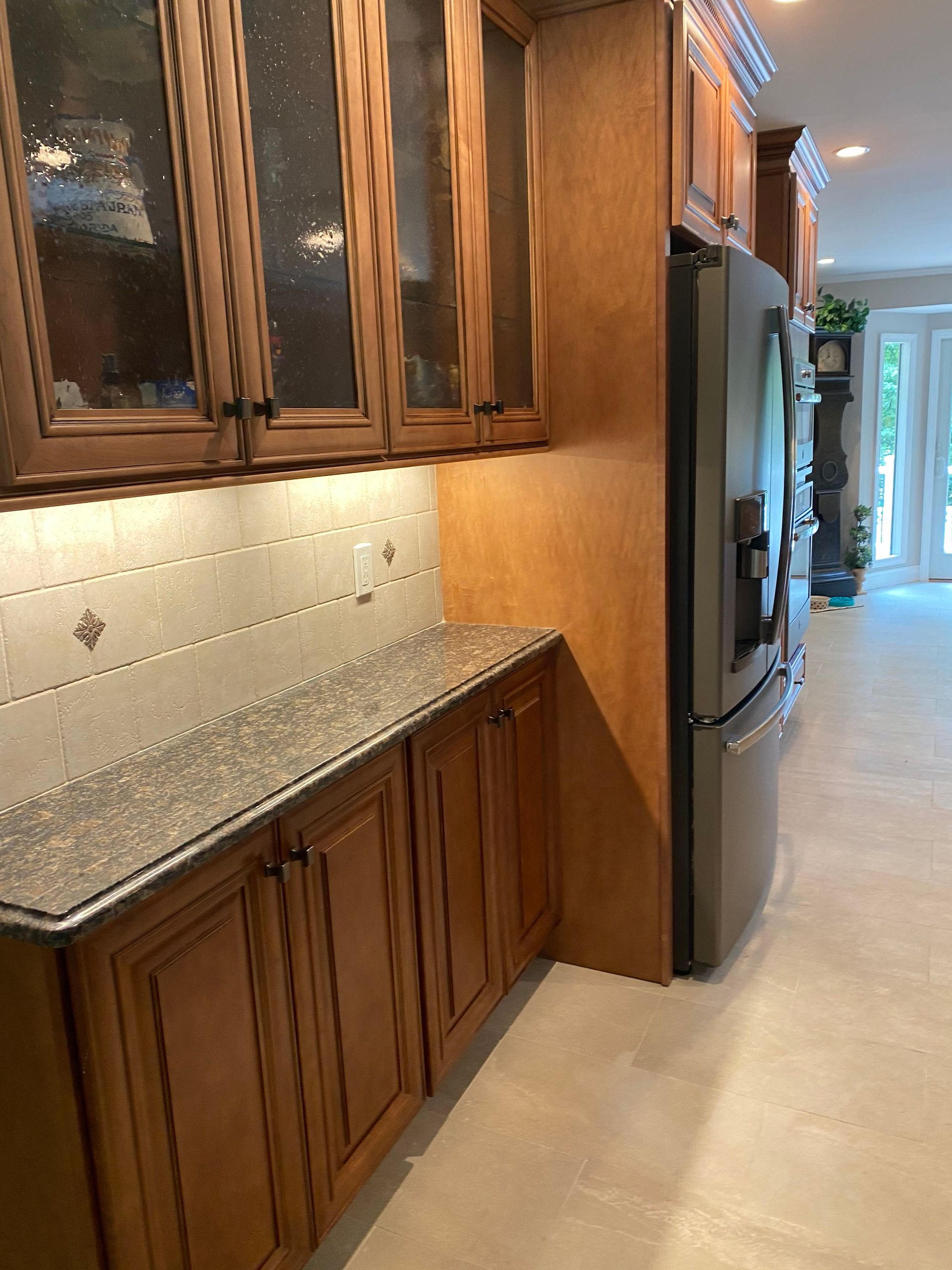 Kitchen hallway with wooden cabinets, granite countertop, stainless fridge, and tiled floor.