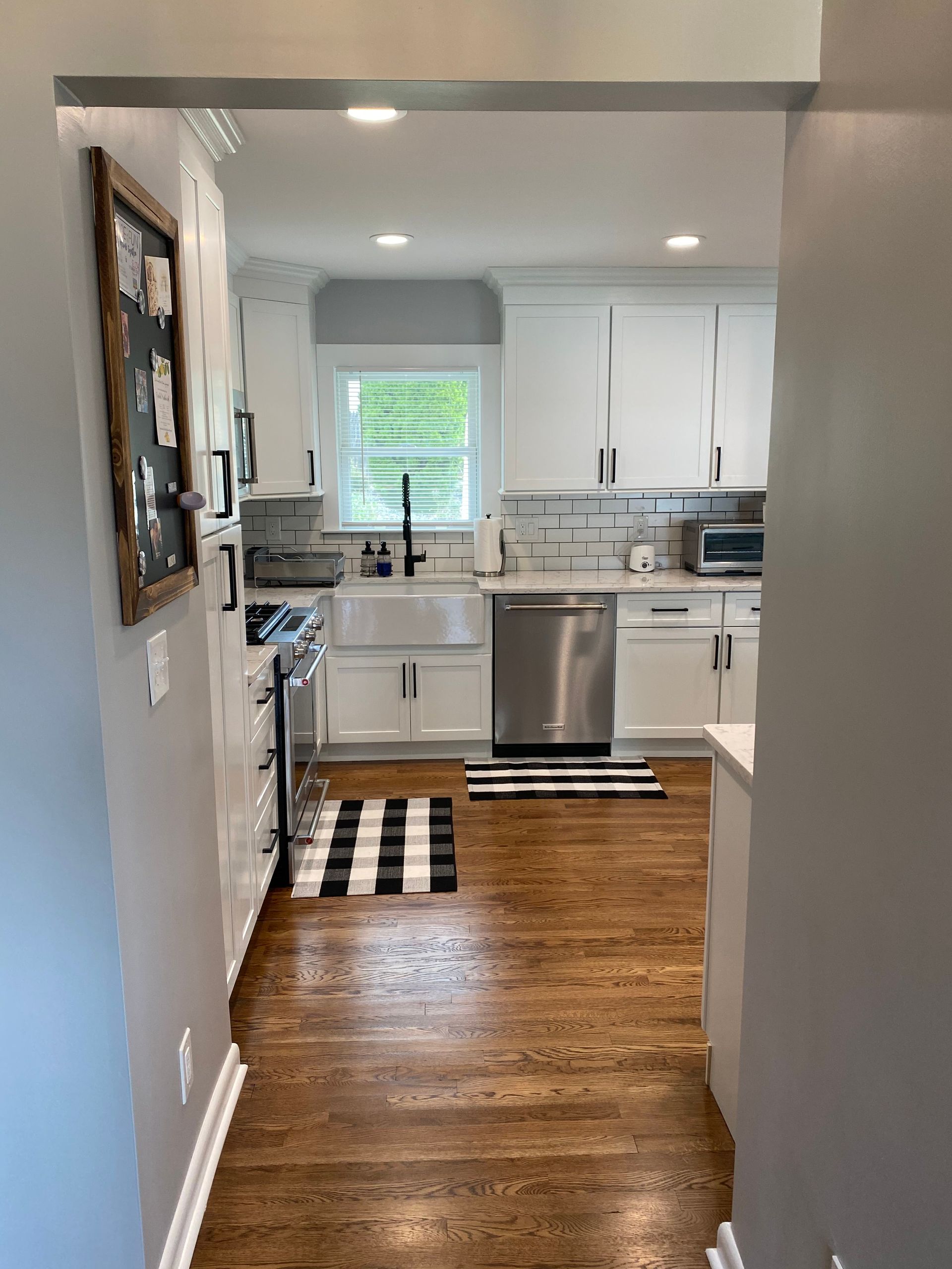 Bright kitchen with white cabinets, stainless dishwasher, checkered rug, and a window over the sink.