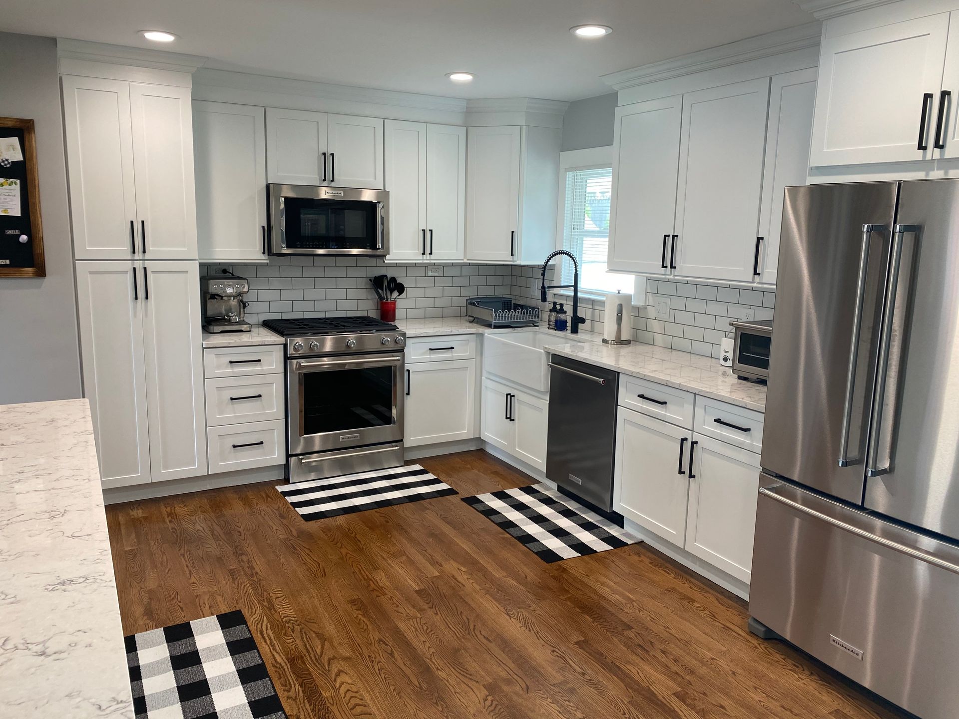 Bright white kitchen with stainless steel appliances, hardwood floor, and black-and-white rugs.