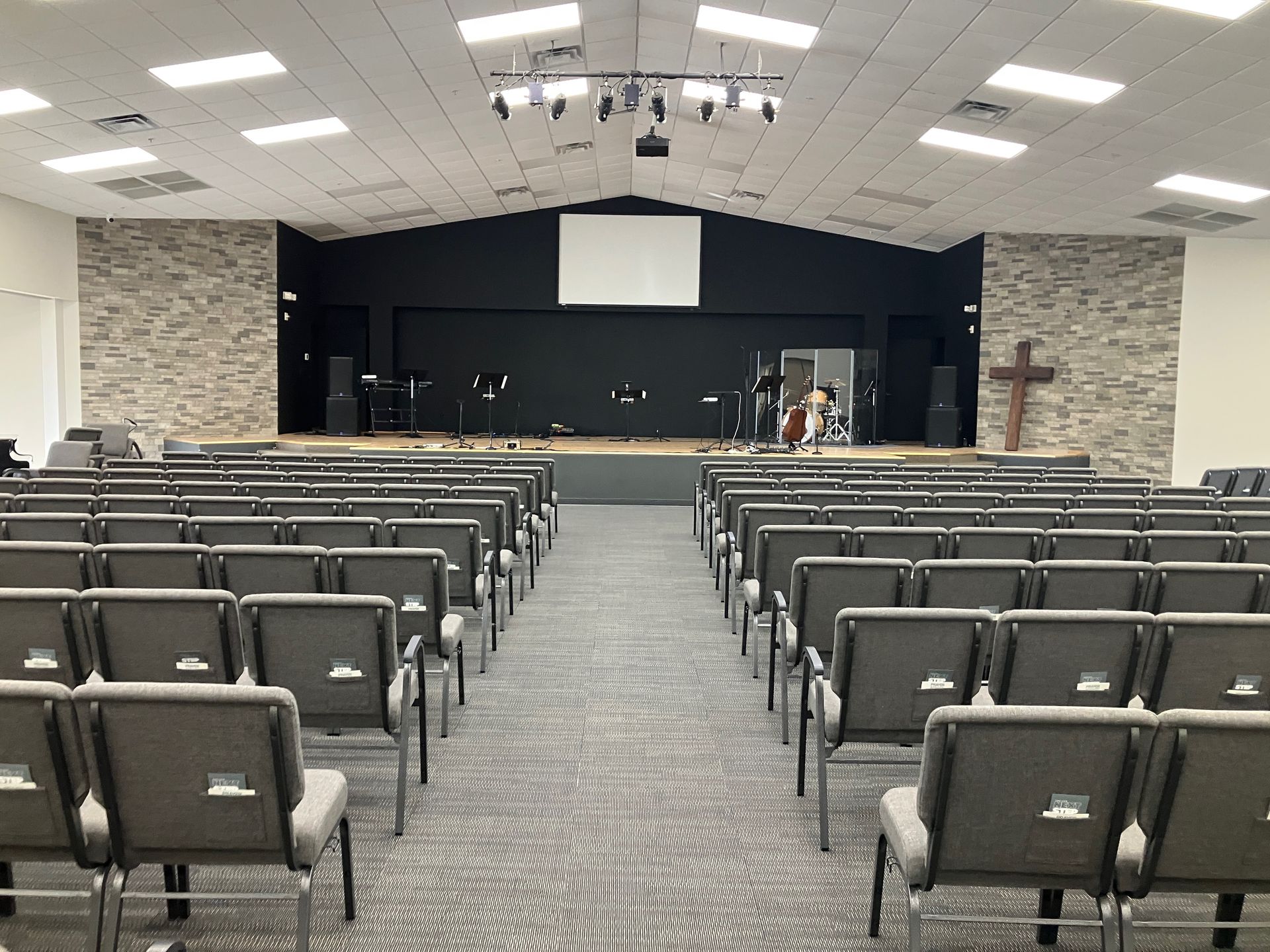 Empty church sanctuary with rows of gray chairs facing a stage and cross under bright ceiling lights