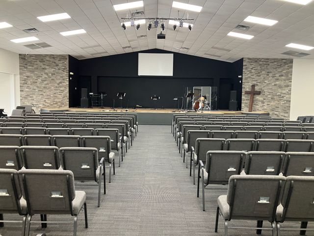 Empty church sanctuary with rows of gray chairs facing a stage and cross on the right.