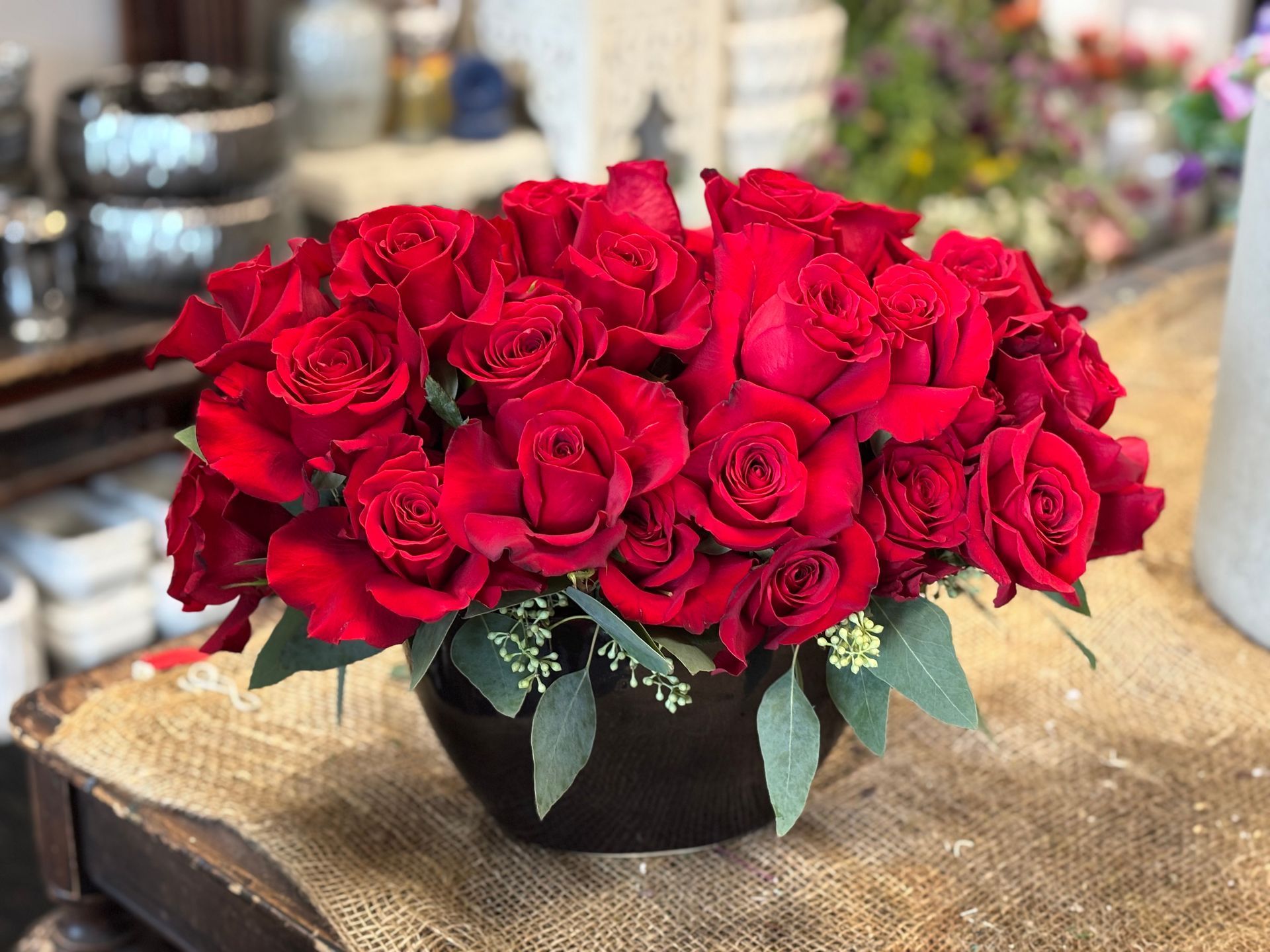 Arrangement of red roses in a black, round ceramic vase, accented with green foliage, on a wooden surface