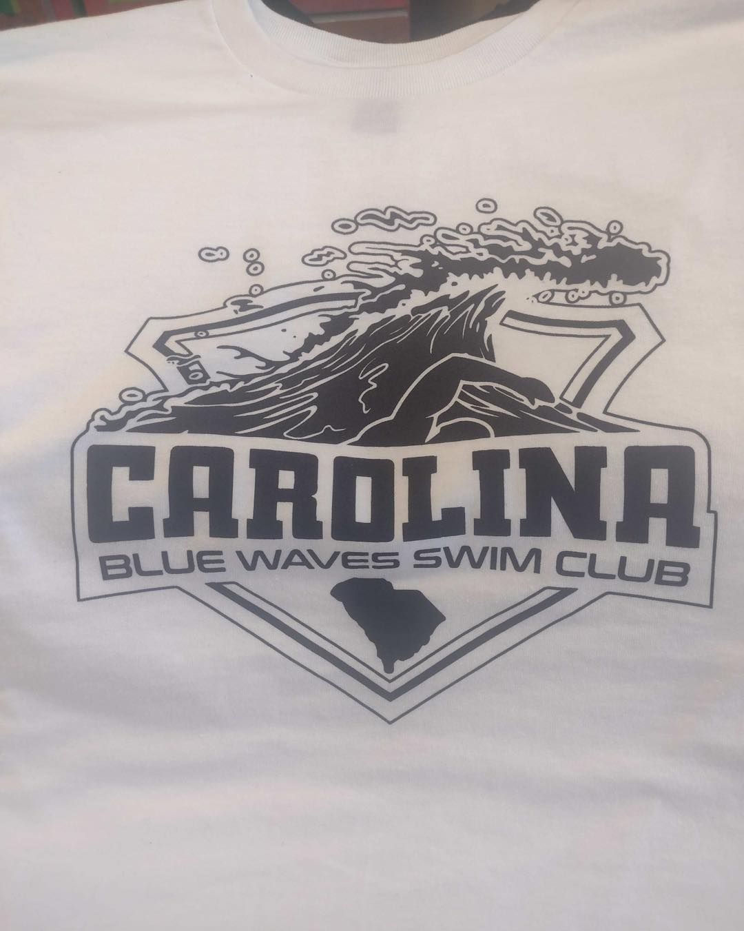 White t-shirt with Carolina Blue Waves Swim Club logo: wave graphic, text, SC outline.