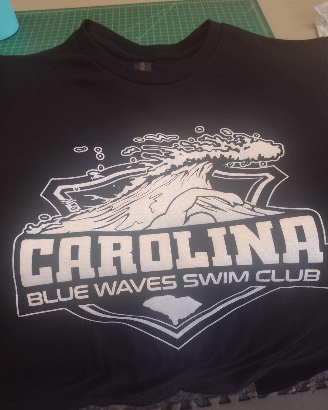 Black t-shirt with Carolina Blue Waves Swim Club logo, a white wave graphic within a shield.