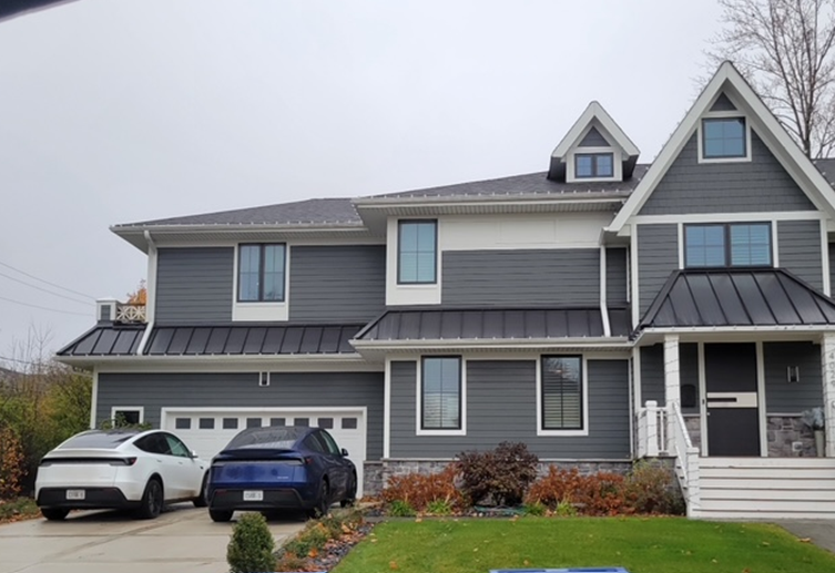 Gray house with black roof, two electric cars in the driveway. Overcast sky.