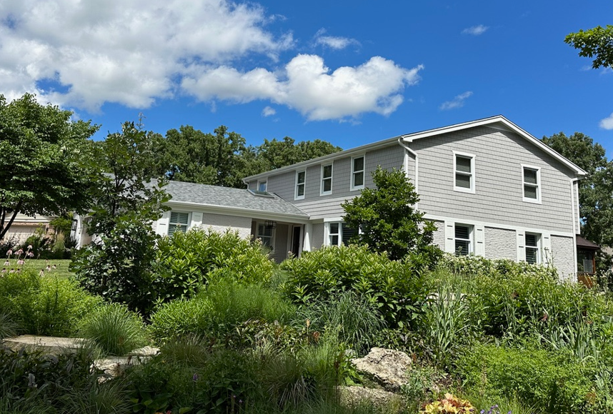 A large white house is surrounded by trees and bushes on a sunny day.