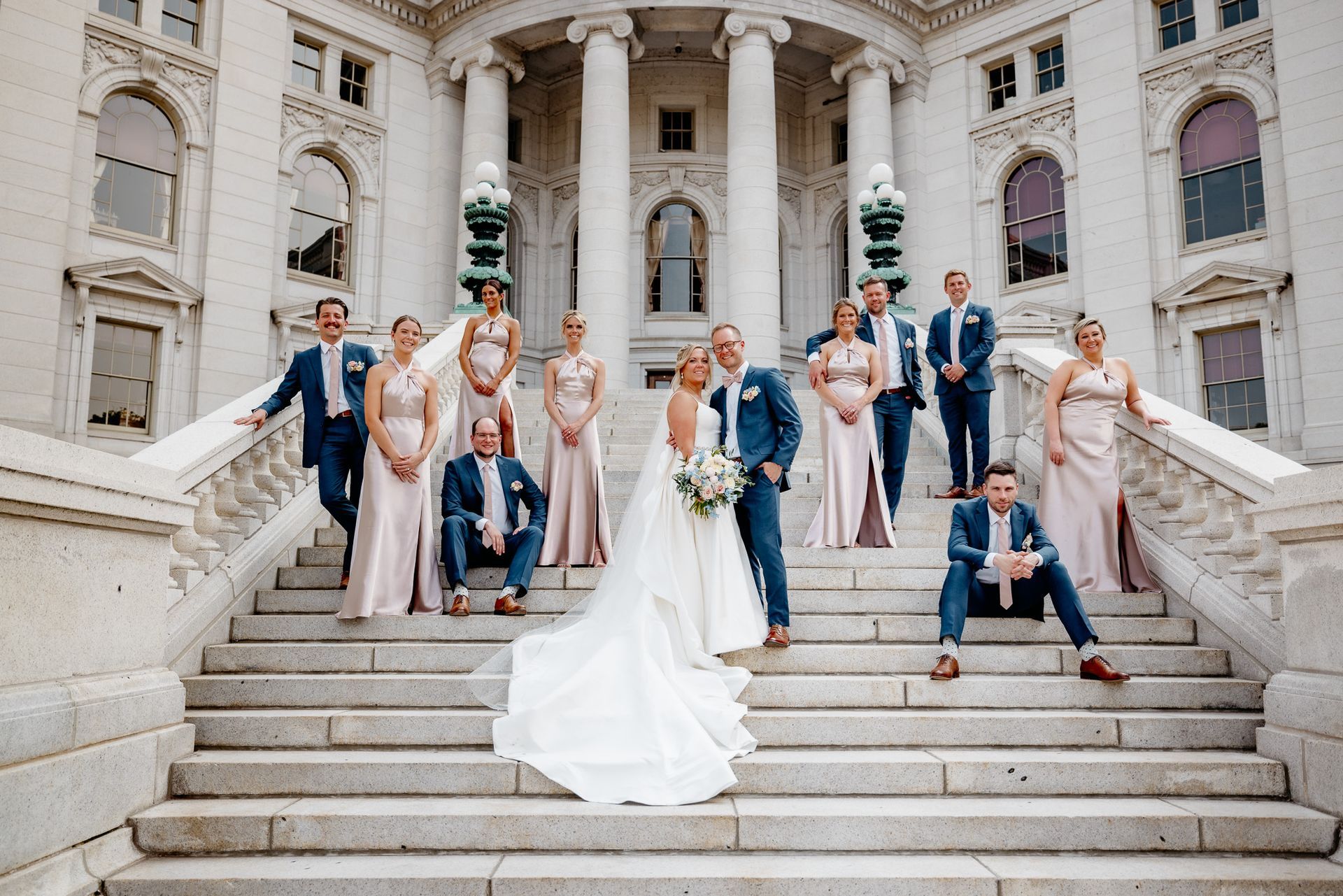 A bride and groom are posing for a picture with their wedding party on the steps of a building