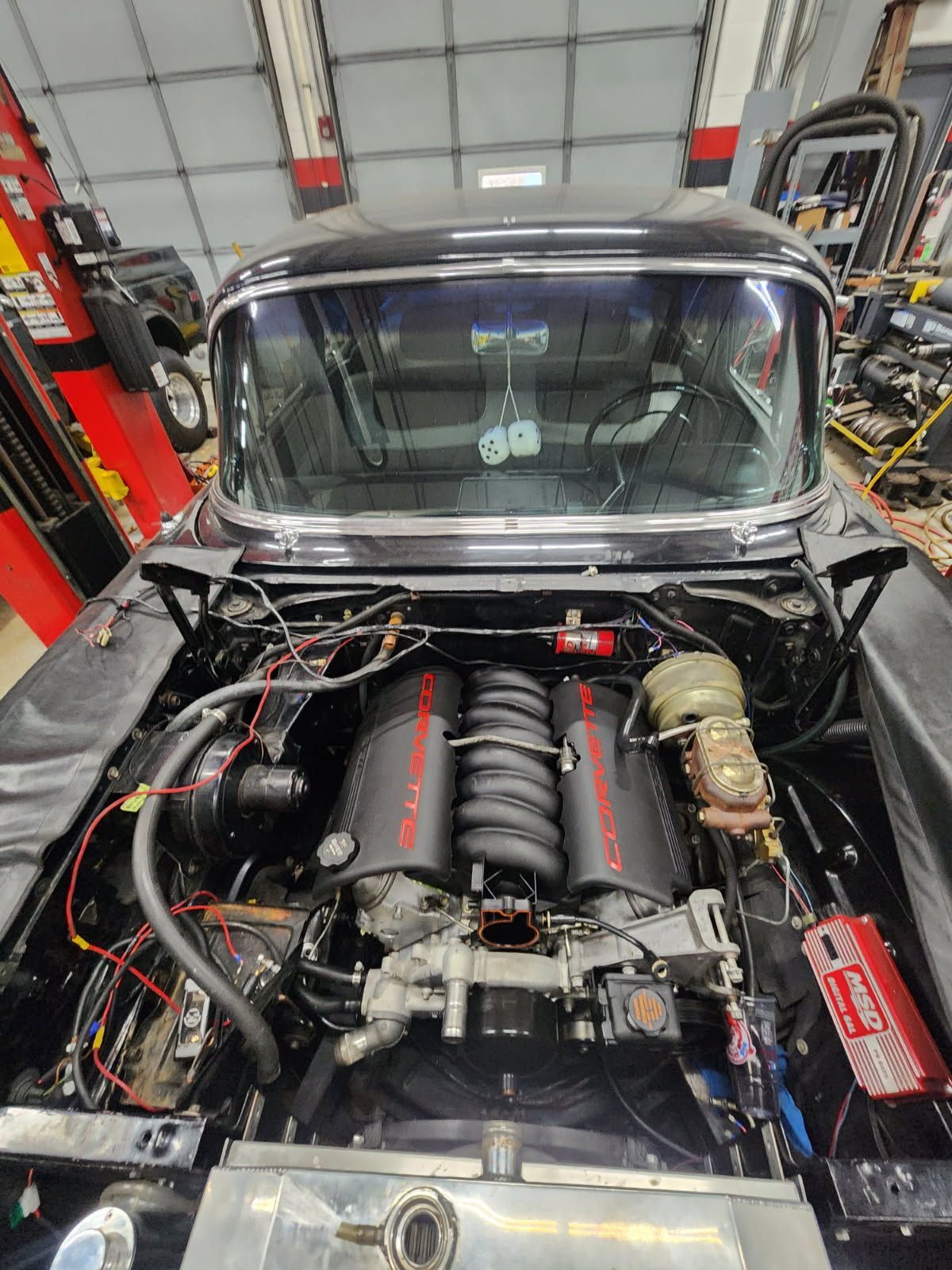 Classic car engine bay in a garage, with a large V8 engine and exposed wiring.